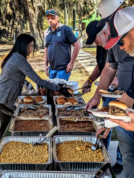 Guests serve themselves from a buffet of macaroni, beans, and burgers at an outdoor gathering, showcasing a social and festive atmosphere.