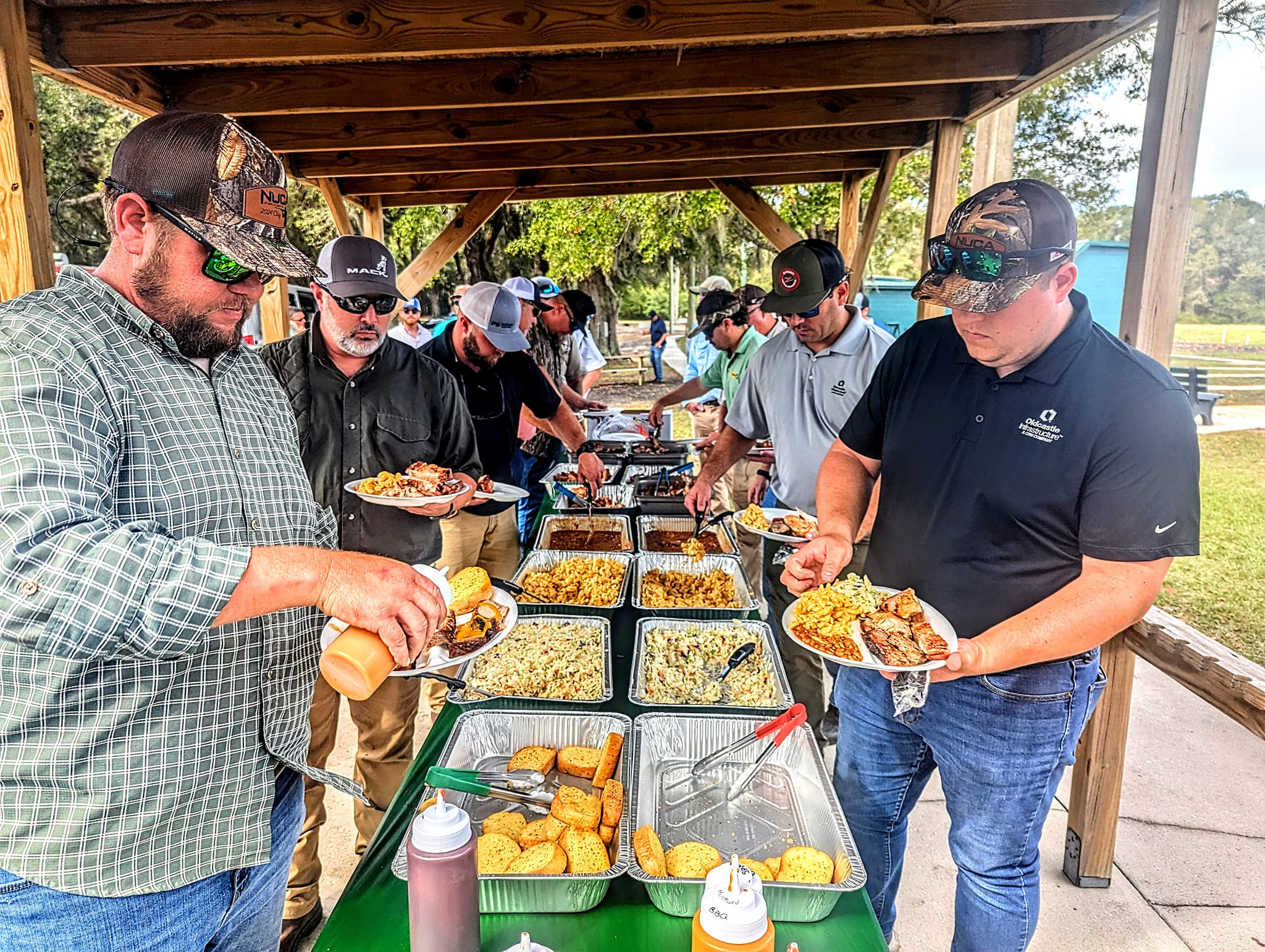 A group of men at a picnic pavilion serve themselves from a buffet of barbecue dishes, including meat, side dishes, and bread, enjoying a meal together.