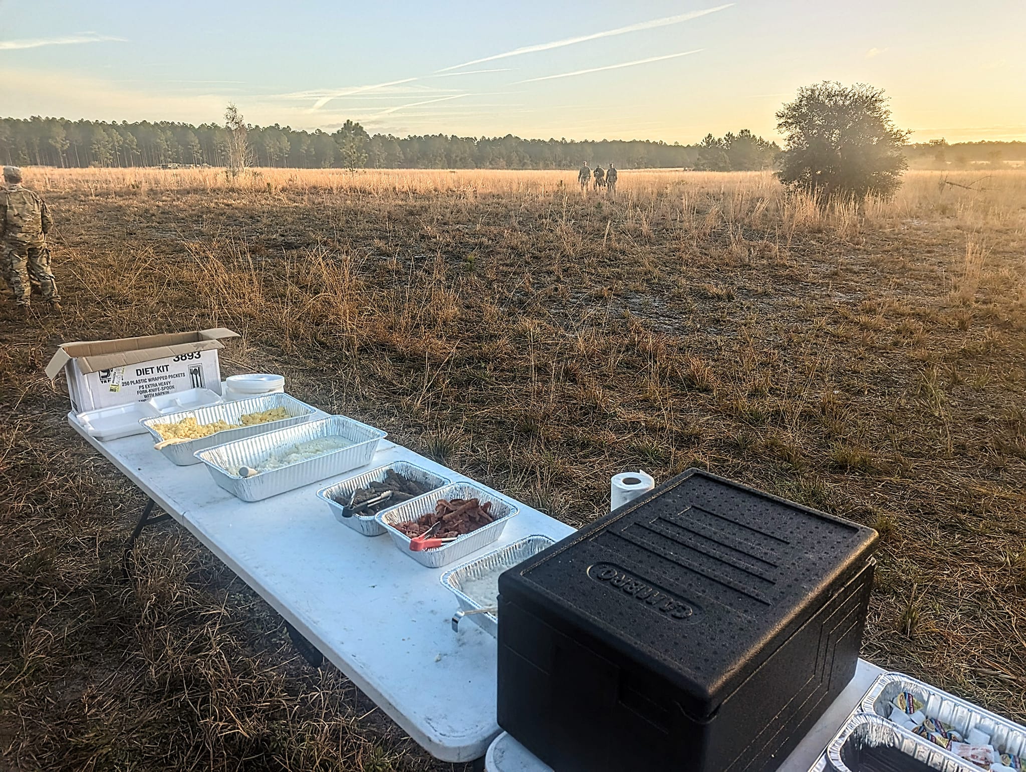 A table displays various food containers in a grassy field during sunrise, with soldiers walking in the background, highlighting a military meal setup.
