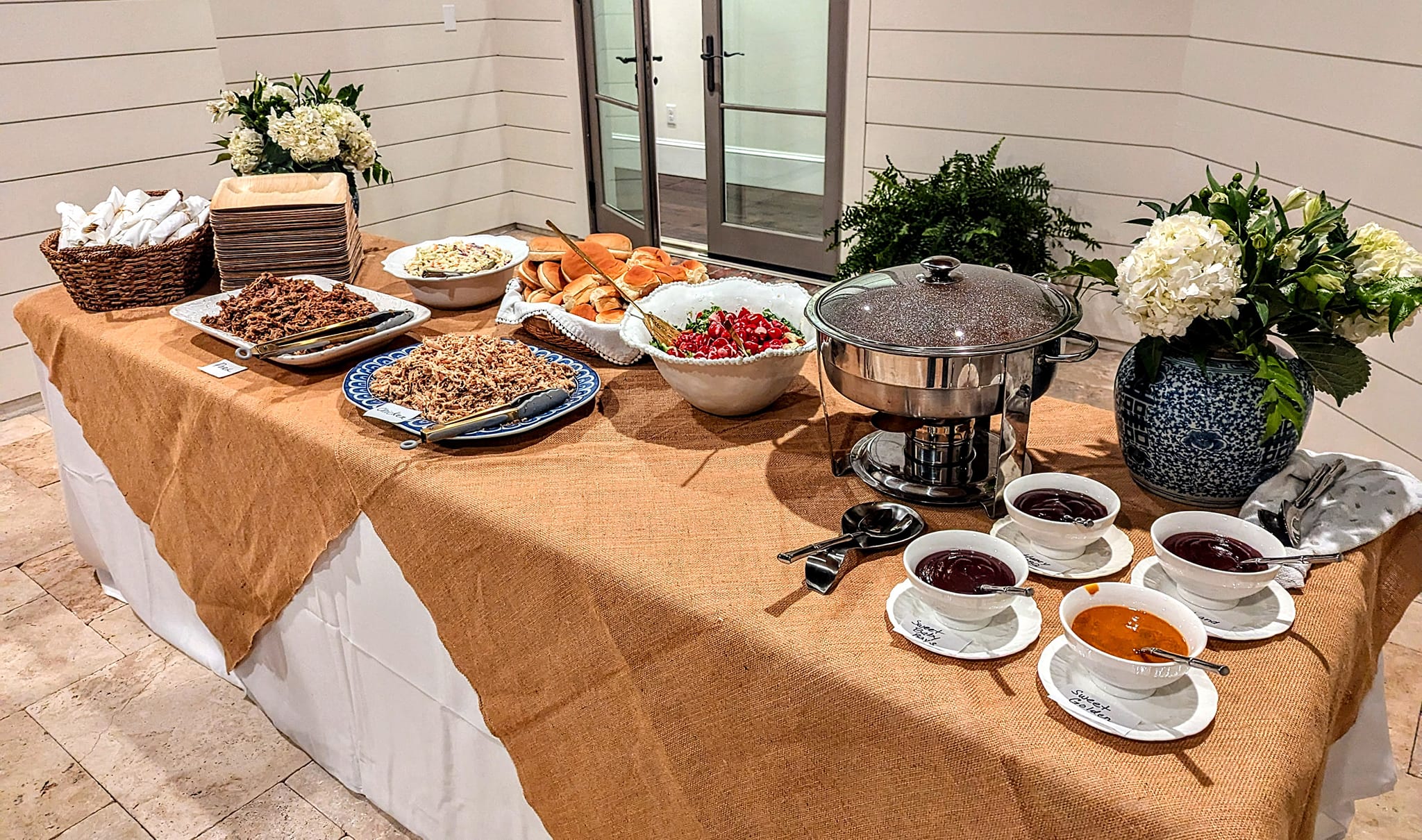 A banquet table featuring various dishes: pulled meats, salads, rolls, sauces, and floral decor, ideal for a catered event or gathering.